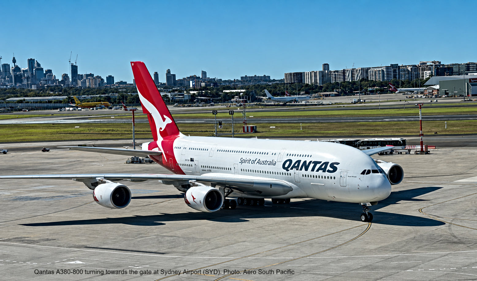 A Qantas Airbus A380-800 turning on the tarmac at Sydney Airport, with the red kangaroo logo on its tail and the Qantas name on the fuselage. The background shows the Sydney city skyline under a clear blue sky.